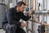 Plombier qualifié écoutant une canalisation et consultant un manomètre dans un local technique sec.