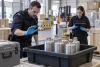 Deux agents des douanes inspectent des flacons métalliques scellés dans un bac de rétention.